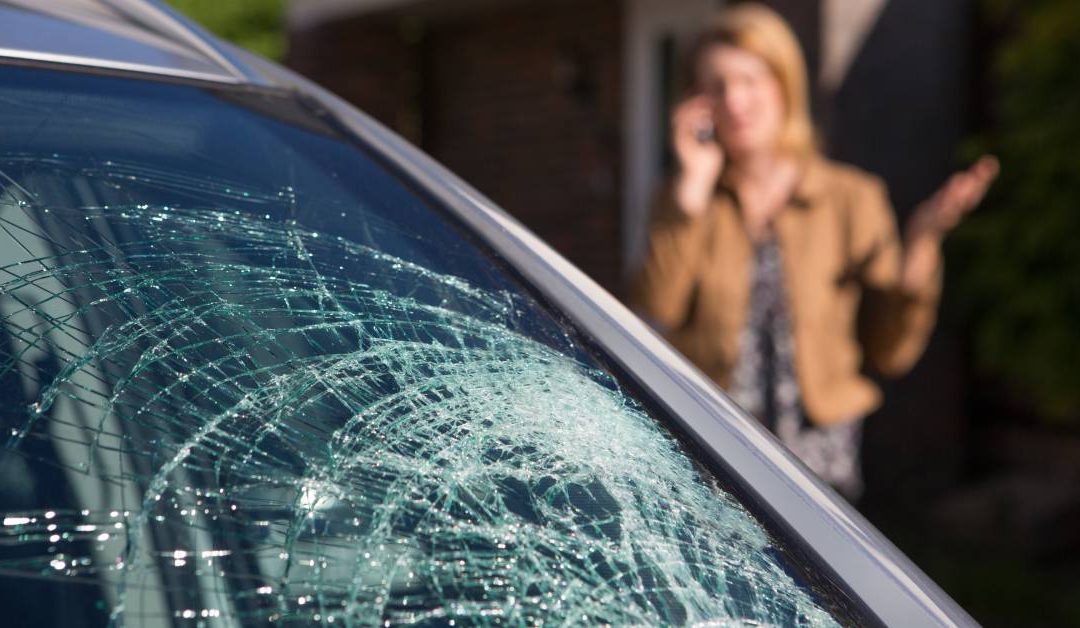 A woman talking on her phone while looking at a cracked car windshield and making an annoyed expression.