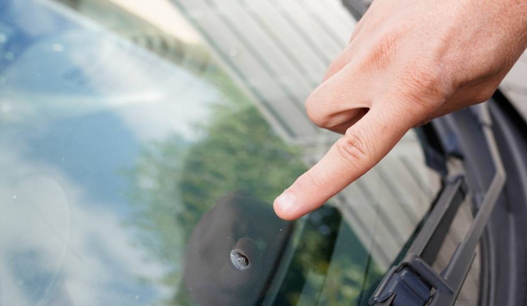 A person’s index finger pointing at small divot in a car windshield. The sky and trees are reflecting off of the glass.