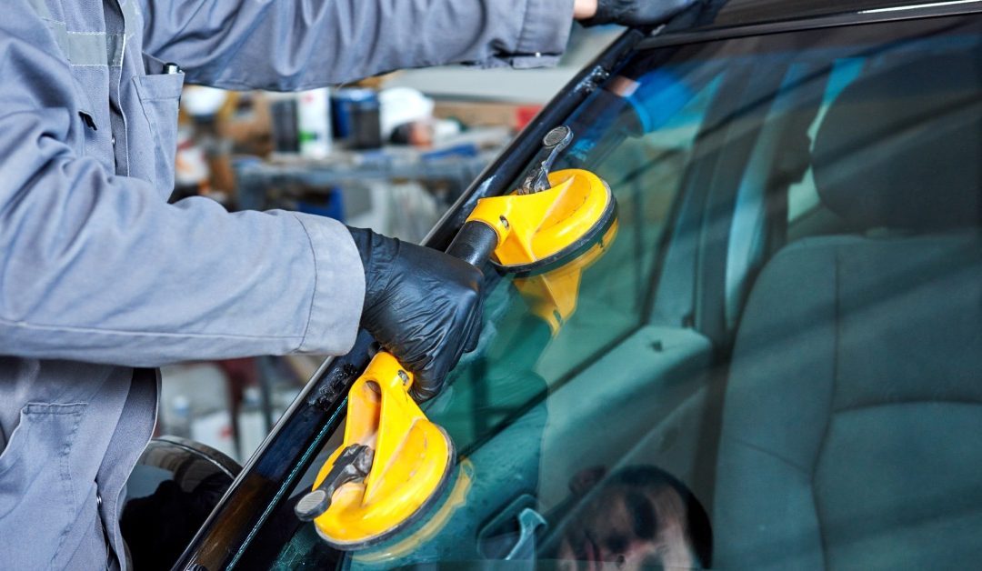 An automotive worker wearing rubber gloves and a long-sleeve jacket replacing a windshield on a car with a yellow tool.