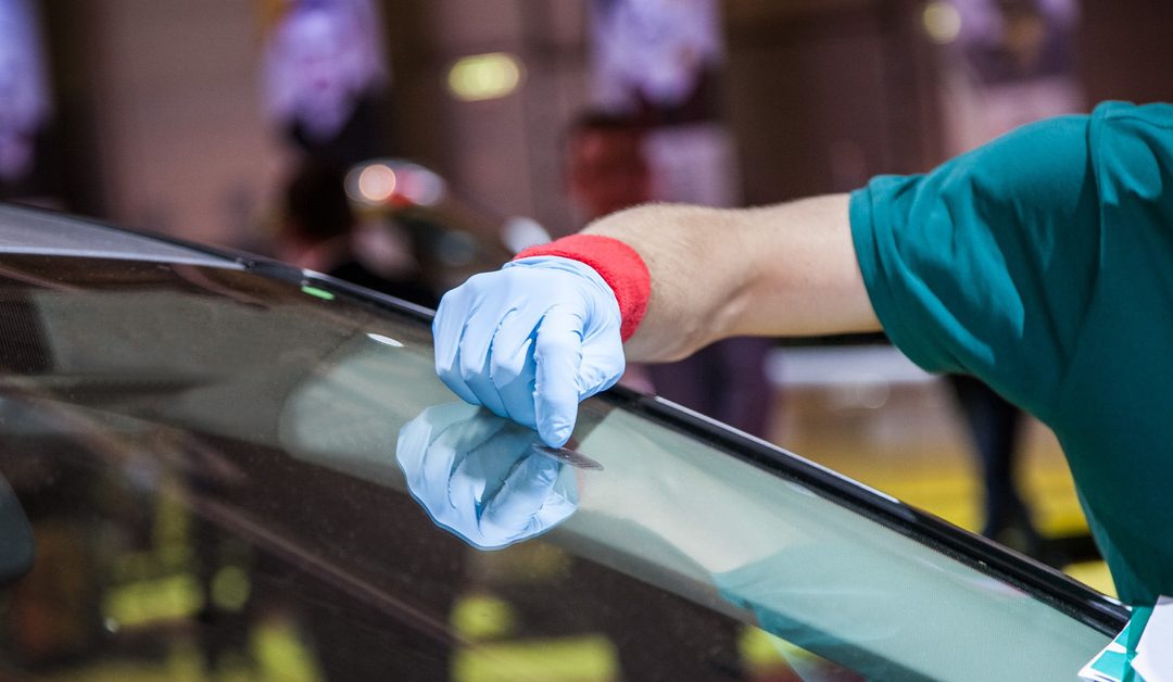 A repair technician wearing a green work shirt and blue rubber gloves touching a crack in a windshield.