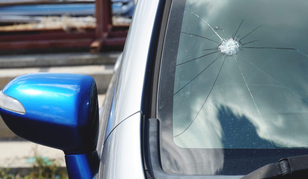 A silver car with a blue side-view mirror. There is a large starburst crack in the windshield with cracks radiating from it.