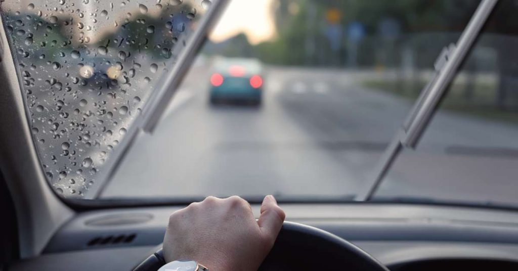 A person’s hand is on a steering wheel. Through the windshield, we see it's raining and the windshield wipers are on.