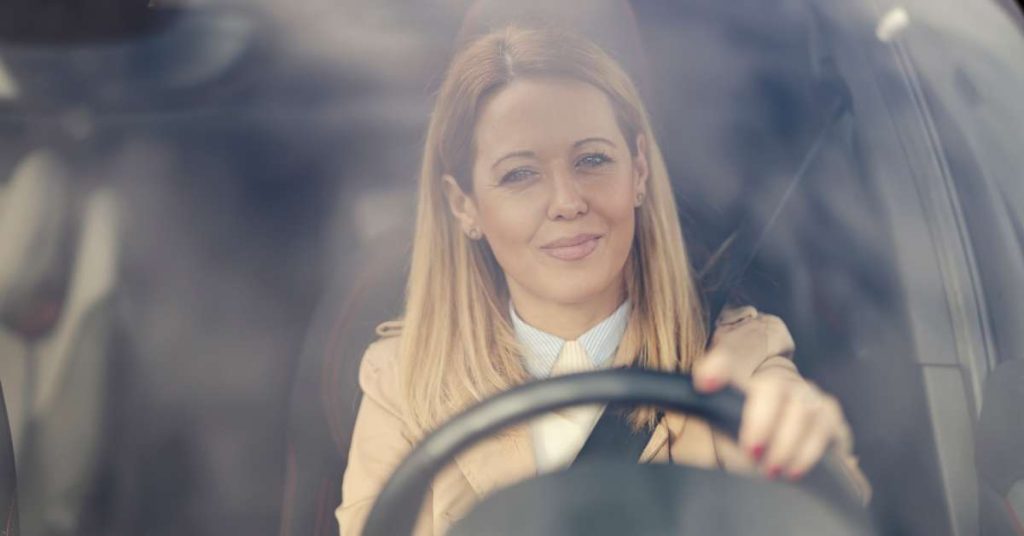 A woman wearing a tan suit sitting in her car looking out through her windshield with a smile on her face.