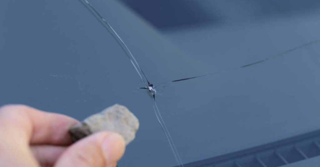 A person’s hand holding a small rock in front of a damaged windshield with a long crack down the center.