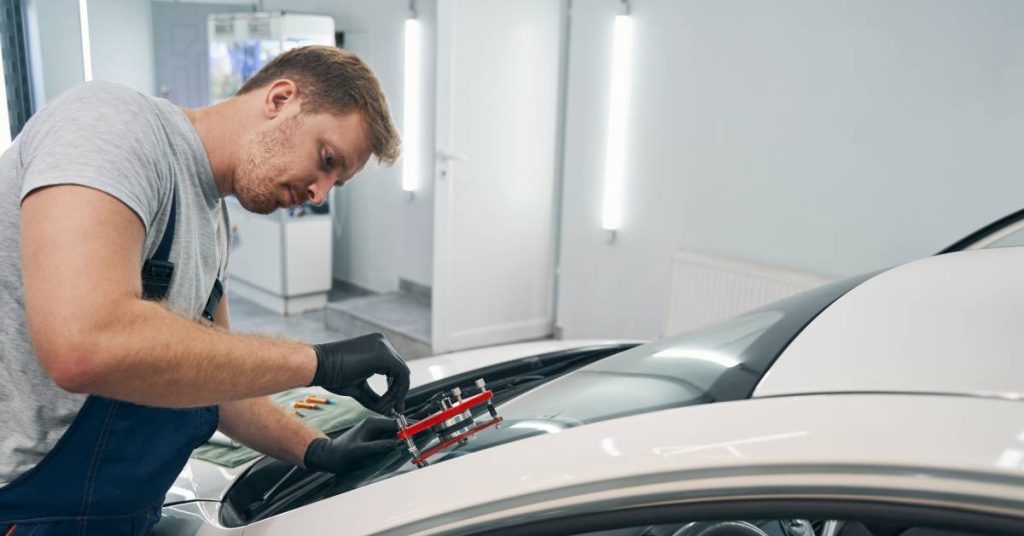 A professional technician wearing gloves and overalls in a shop using a red tool to repair a broken windshield on a white car.