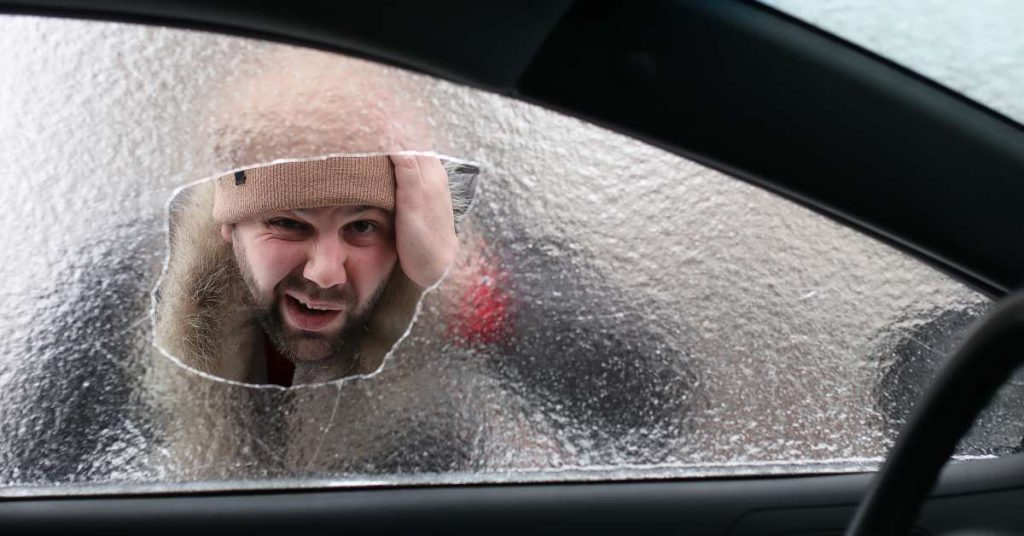 A bearded man in a brown hat looks worried as he examines a large crack in the ice-covered windshield of his car.