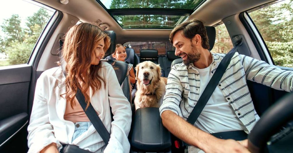 Two adults, a small child and a golden retriever dog smile in a four-door car with trees in the background.