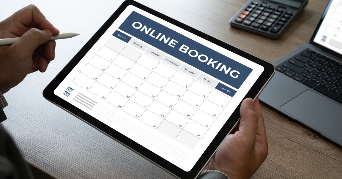 A wooden desk with a calculator and a laptop. A person holds a tablet that reads, "ONLINE BOOKING," above a calendar.