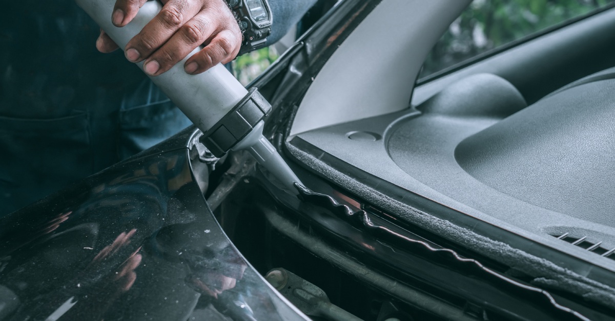 A black vehicle's car cowl at close range. A person uses a cylindrical tool to apply an adhesive to the cowl's edge.