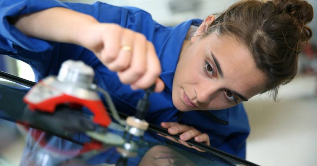 A female mechanic in a blue shirt concentrates as she uses professional tools to repair a car's windshield.