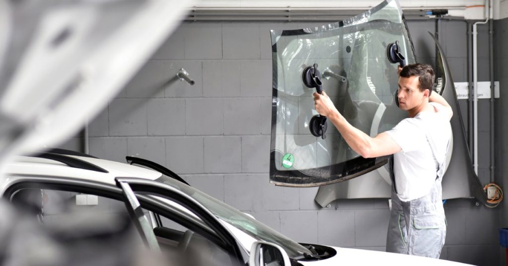 A mechanic in overalls and a white T-shirt prepares to replace the windshield on a small silver car.