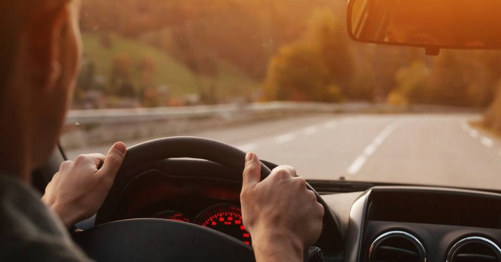 An over-the-shoulder view of a man holding the steering wheel of a vehicle as he drives while the sun is setting.