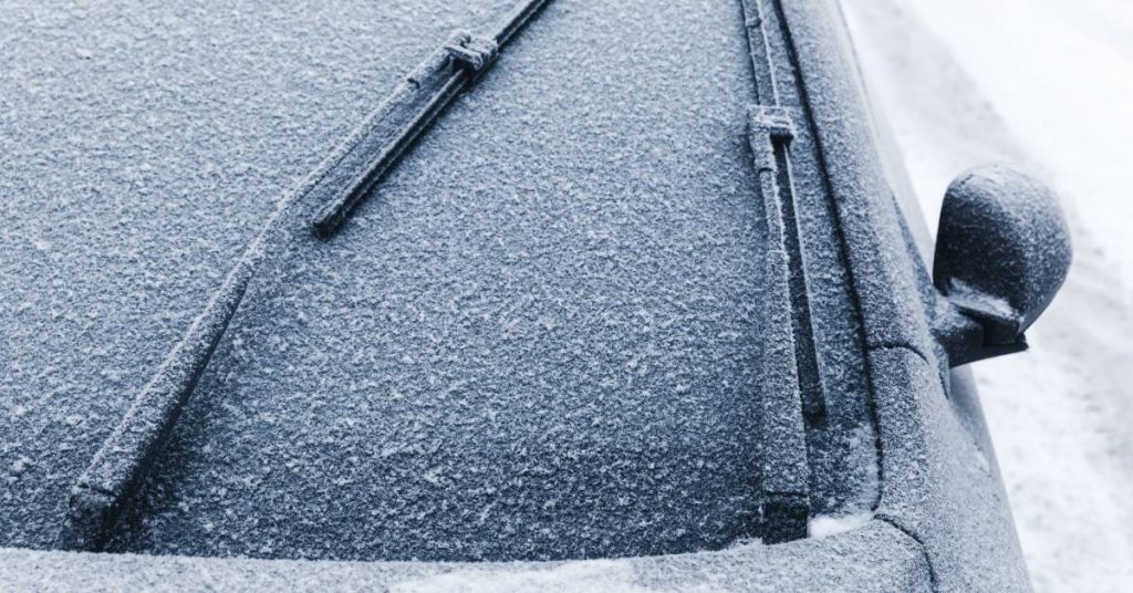 A small, dark colored vehicle is covered in a thin layer of snow. The windshield wipers are moving.