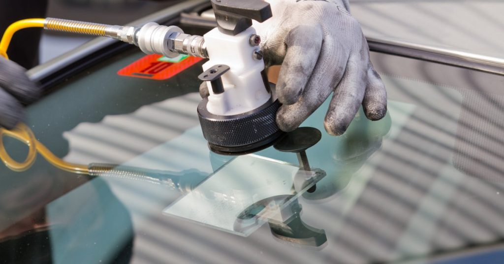 A hand wearing a dirty white work glove is holding a large tool to fix a crack in a car's windshield.