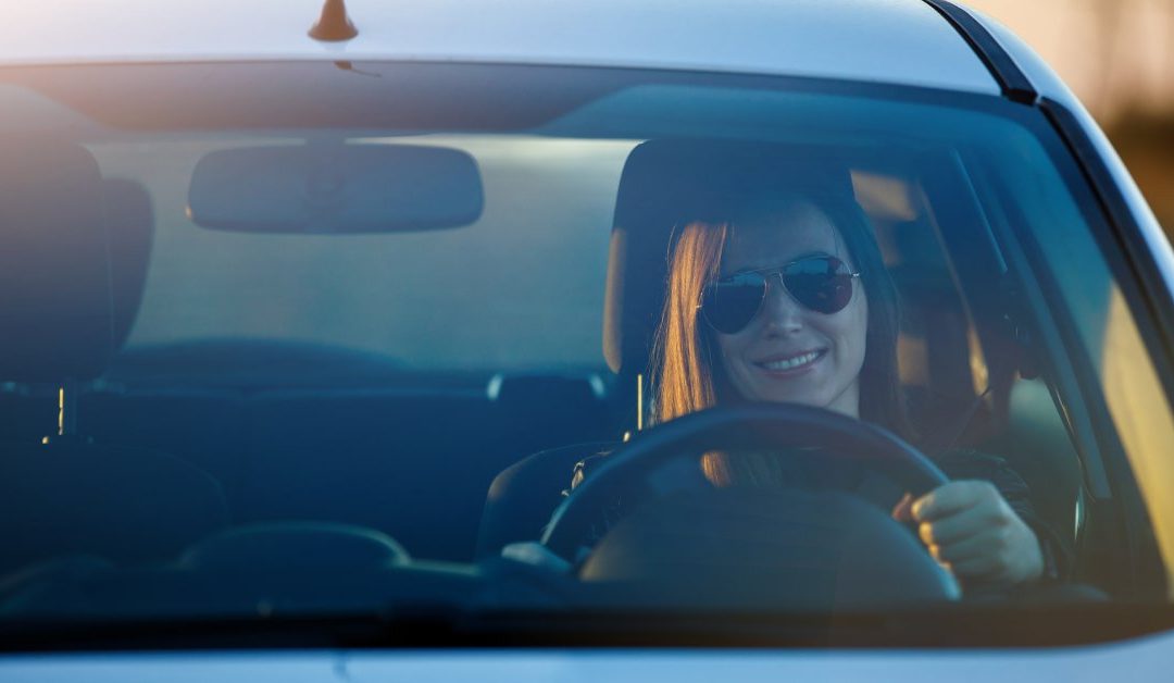 A young woman wearing sunglasses smiles as she drives a small silver vehicle and the sun appears to be setting.