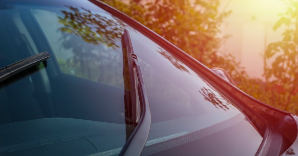 A vehicle's windshield wipers are moving over the windshield. The glass is reflecting the glow of the sunset.