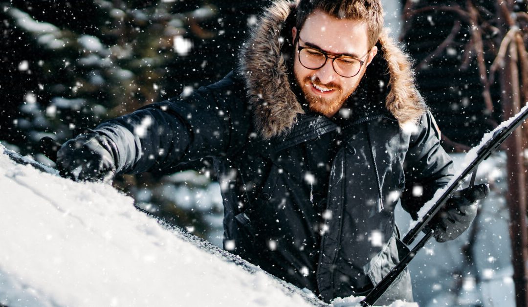 A smiling man wearing a black winter coat cleans snow off his windshield with snow falling all around him.