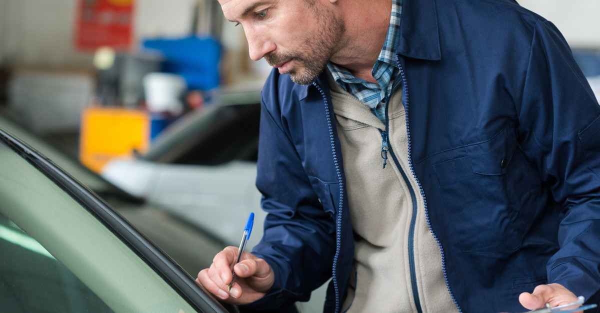 A man wearing a blue jacket and holding a clipboard and pen closely inspects a small vehicle's windshield.