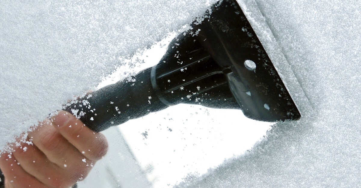 A close-up of a person's hand using a windshield scraper to scrape a layer of snow off a car's windshield.