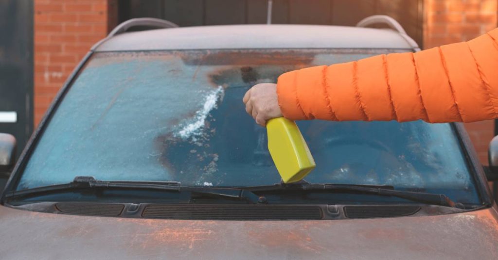 A person wearing an orange coat holds a yellow spray bottle and sprays liquid onto the windshield of a car.