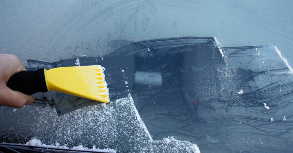 A close-up showing a person using a plastic yellow ice scraper to remove a thin layer of ice from a car's windshield.