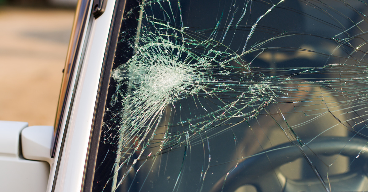 A close-up of a small light-colored vehicle with a massive web of cracks on one side of the windshield.