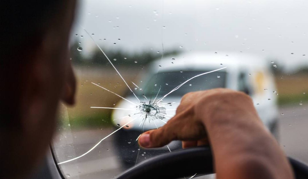 A man drives a car with his right hand on the steering wheel. There is a large crack in the windshield.