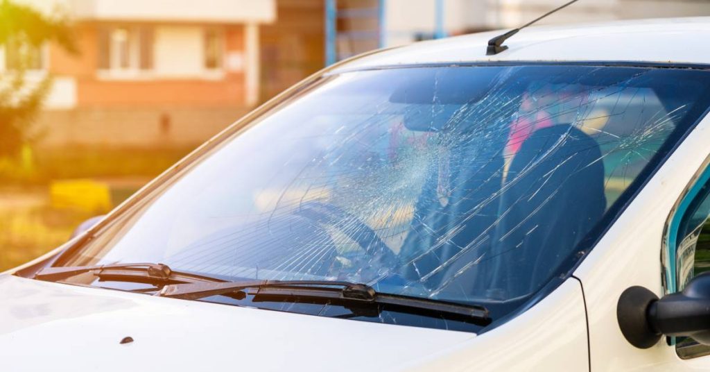 A medium-sized white vehicle with an extremely cracked and shattered windshield sits outside of a house.