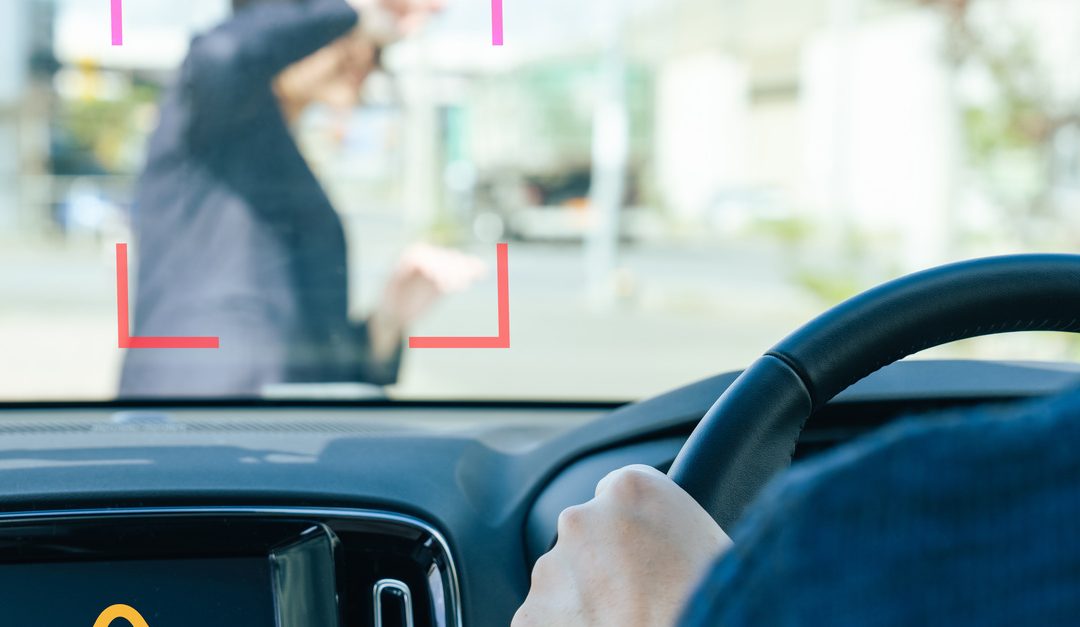 A man is driving a car with a warning symbol displayed on the infotainment screen as someone walks in front of his car.