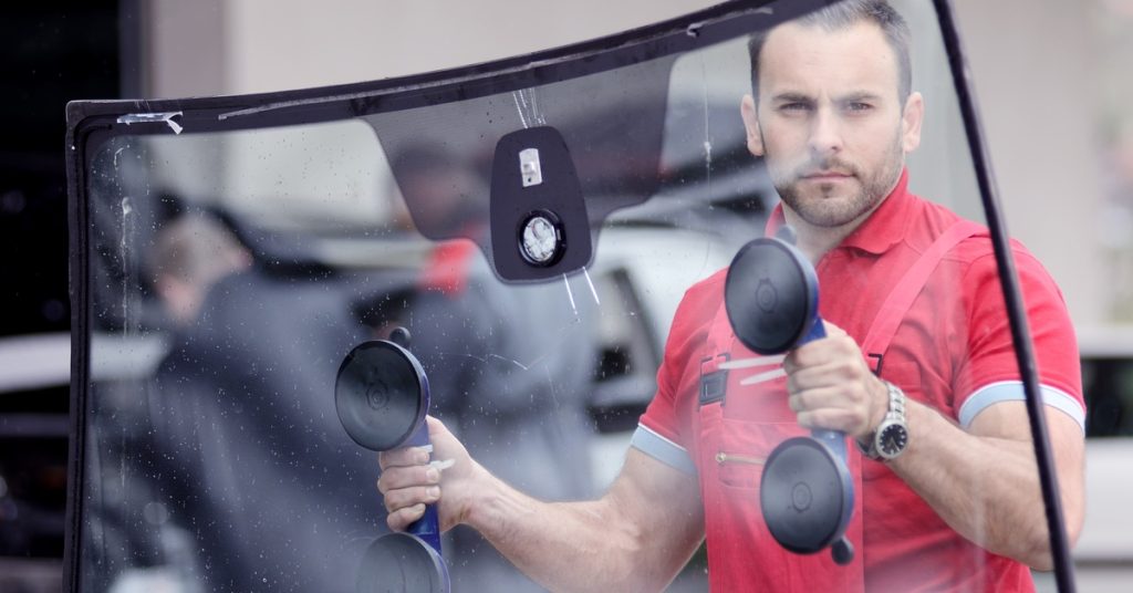 A man wearing a red shirt and apron stands in a garage holding a large windshield with suction handles.