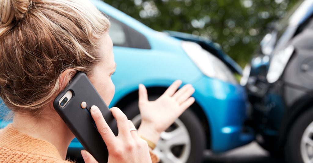 A woman in an orange shirt talks on a cell phone in front of a car accident between one blue and one black car.