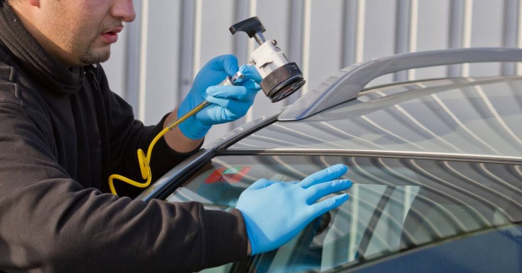 A man wearing a black jacket and blue gloves uses a tool to repair a crack in a silver vehicle's windshield.