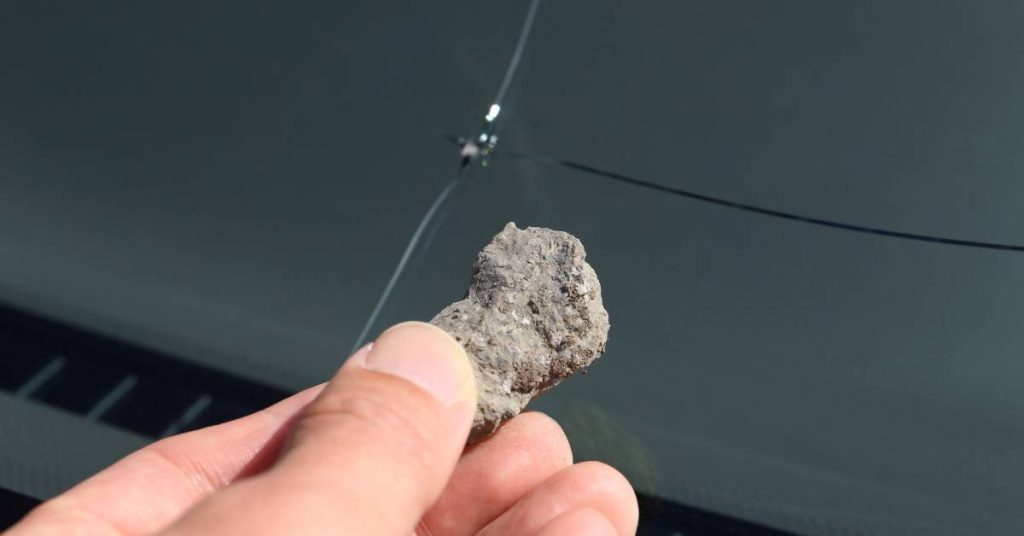 A close-up of a person's hand holding a small gray rock in front of a car windshield with a large crack.