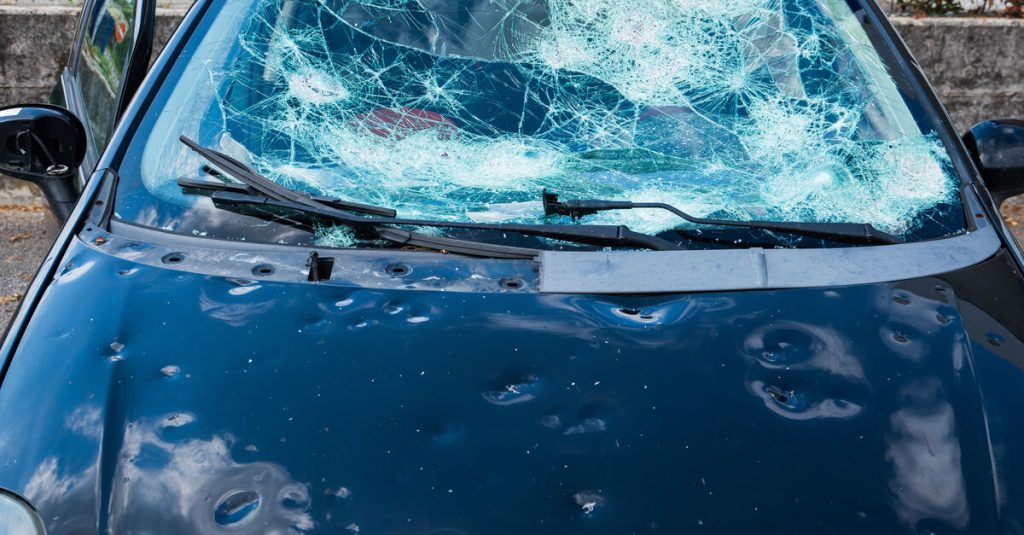 A close-up showing a small black vehicle with small dents scattered across the front and a severely cracked windshield.