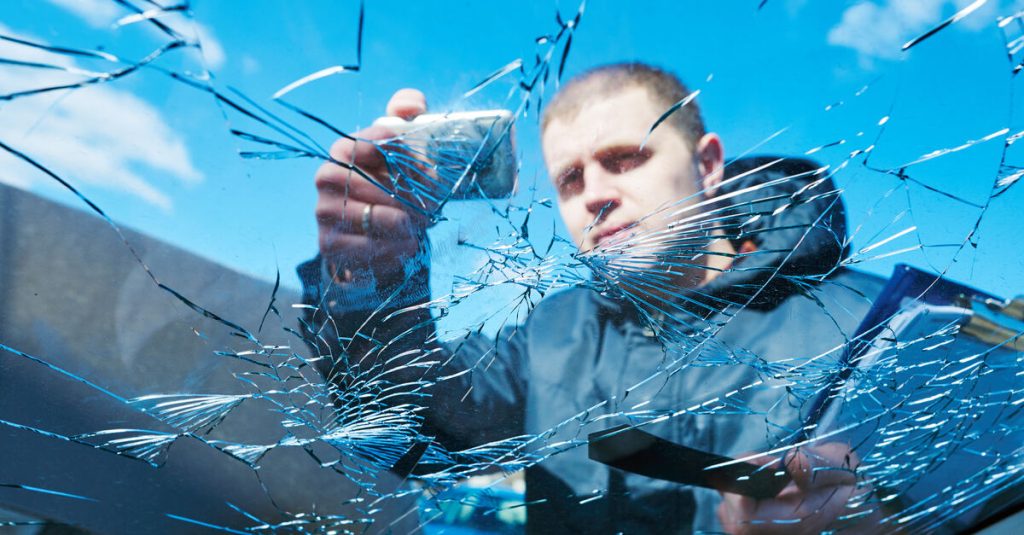 A man wearing a black jacket holds a clipboard as he takes a picture of a windshield with extensive damage.