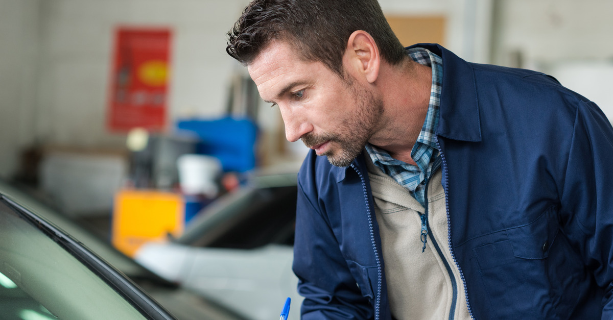 A man in a blue jacket holds a pen and clipboard while closely inspecting the windshield on a small car.