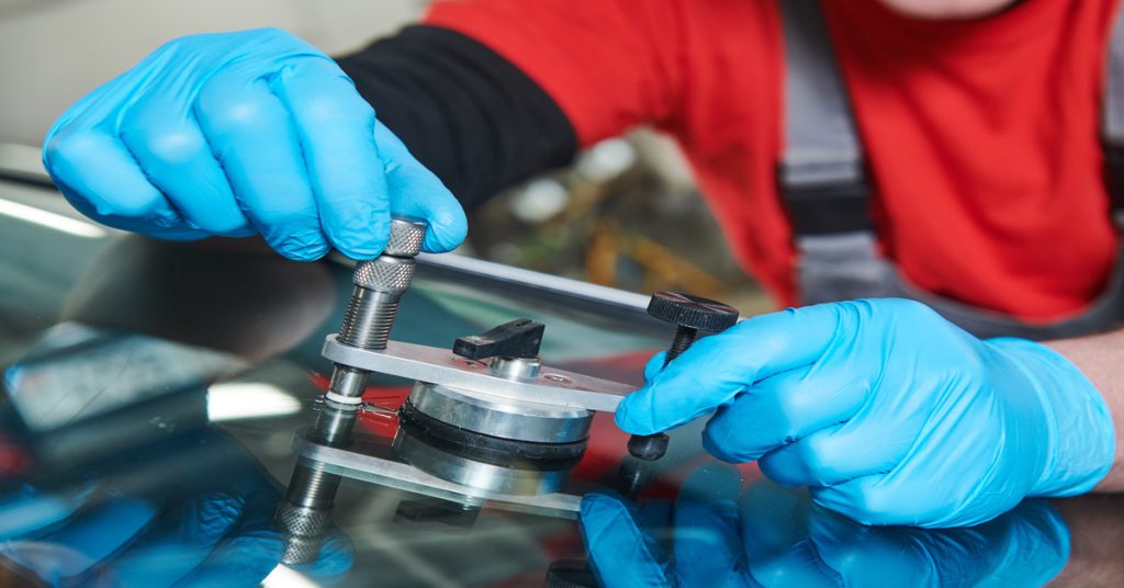 An extreme close-up showing someone wearing blue plastic gloves using a small silver tool to repair a windshield.