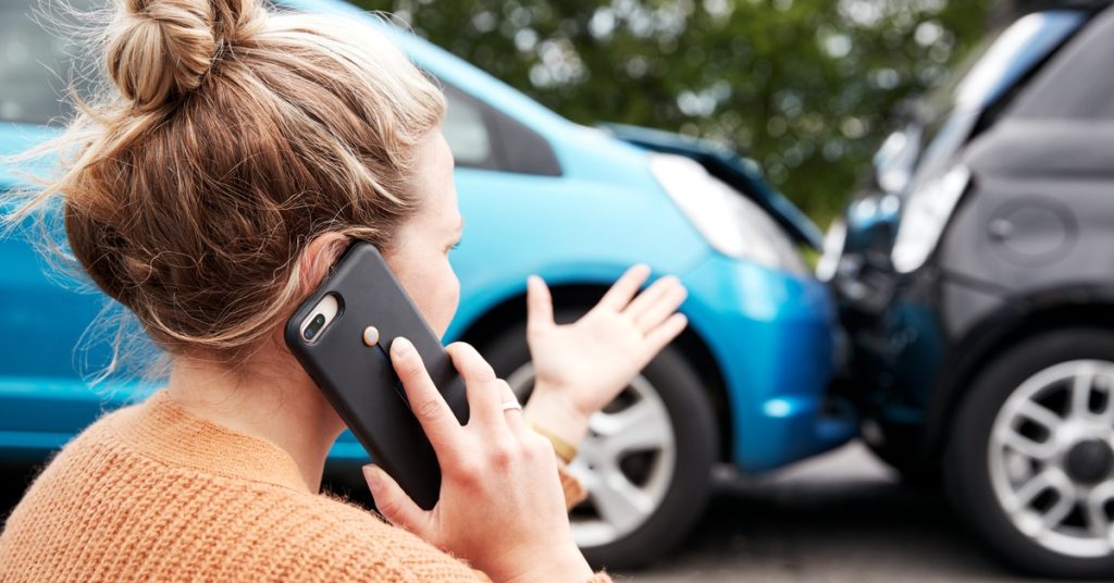 A blonde woman wearing an orange sweater gestures while talking on the phone in front of a car accident.