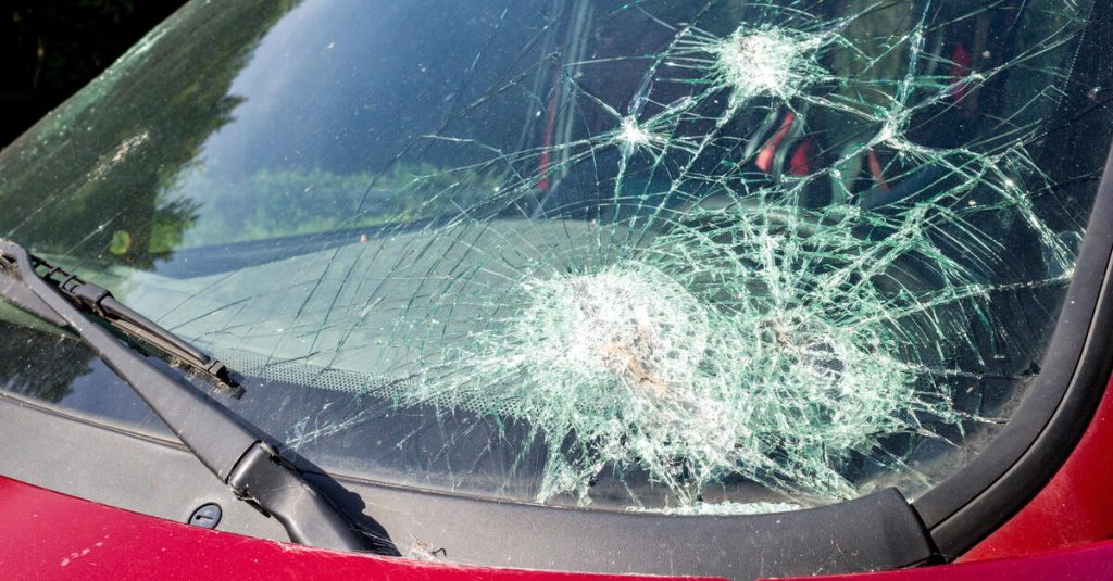 A close-up of a small red vehicle with a very large crack that is spreading throughout the windshield.