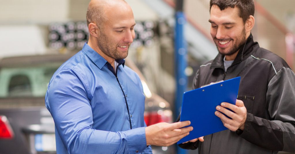 A man wearing a black jacket smiles while holding a blue clipboard up to another man in a blue shirt who is also smiling.