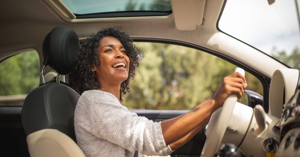 A woman wearing a gray sweater is smiling widely as she drives a car with a tan-colored interior on a sunny day.