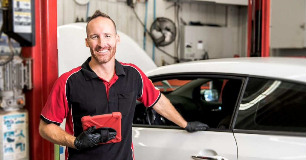 A man wearing a black and red shirt and gloves smiles widely while touching the window of a small, white car.