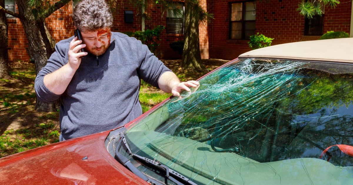 A bearded man in a sweater is talking on a cell phone while examining a very large crack on a small, red vehicle.