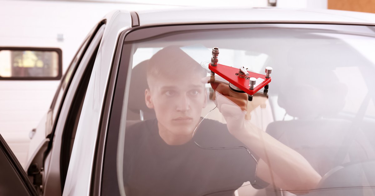 A young man is sitting inside a silver vehicle while using a small red tool to fix a crack in the windshield.