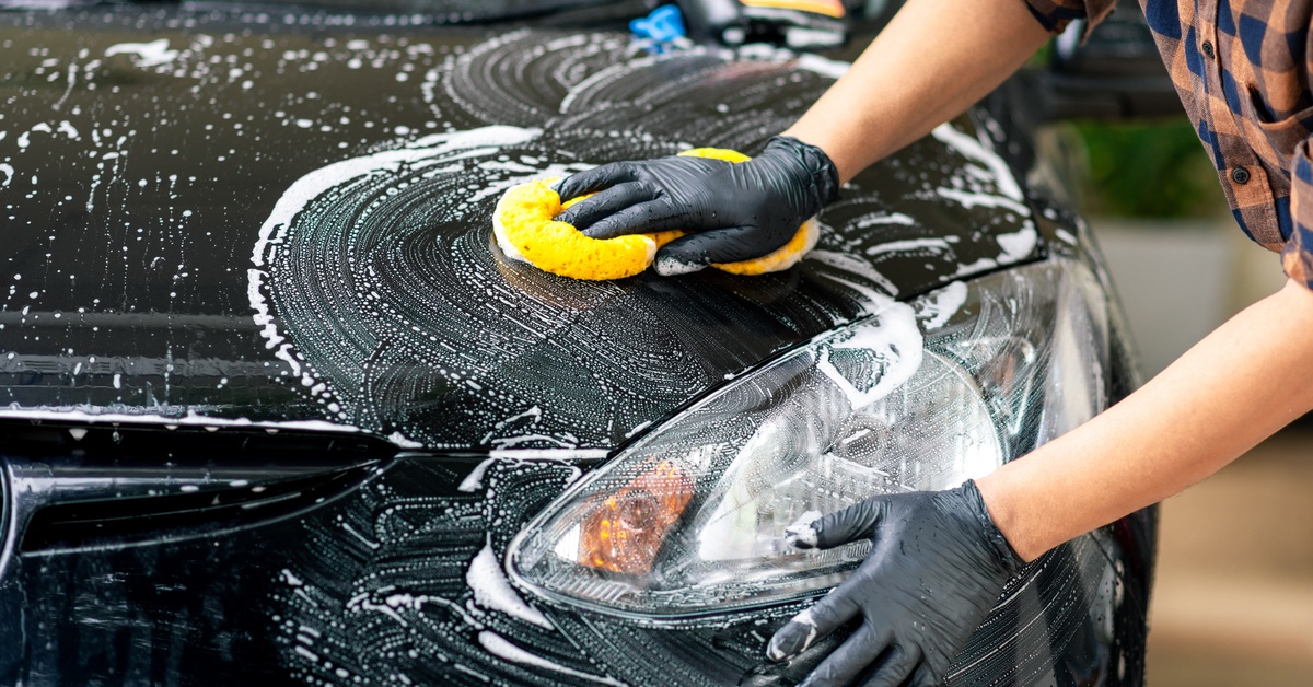 A close-up of a person wearing a plaid shirt and black gloves using a yellow sponge to clean a black car.