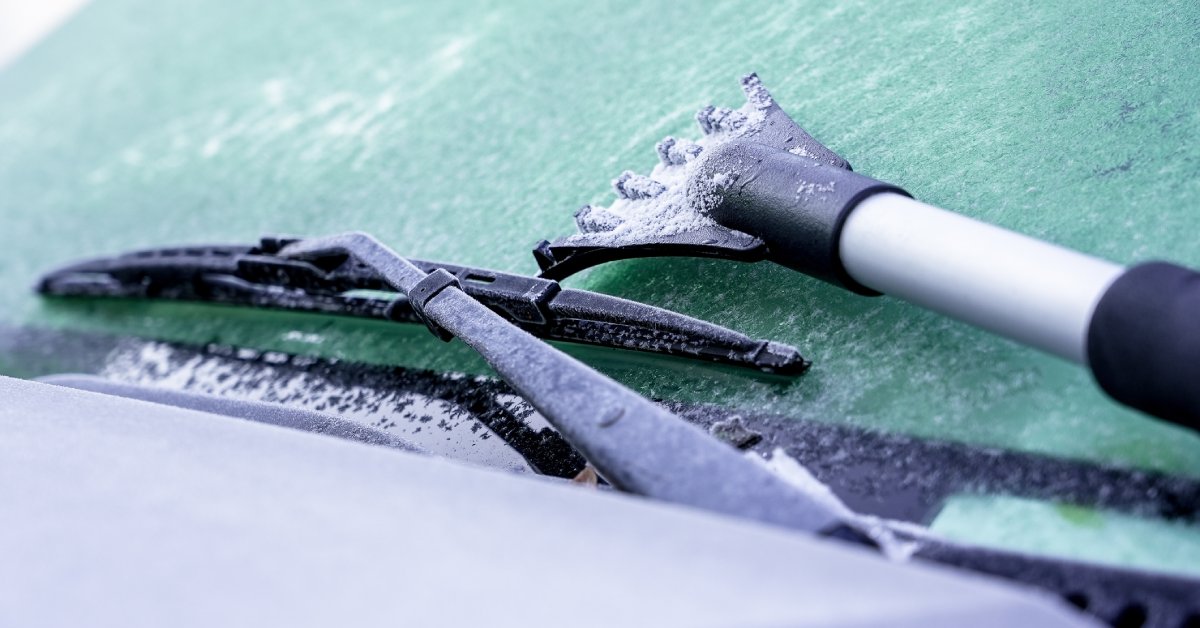 The windshield of a car is covered in frost and ice. An ice scraper is sitting on top of the wipers.