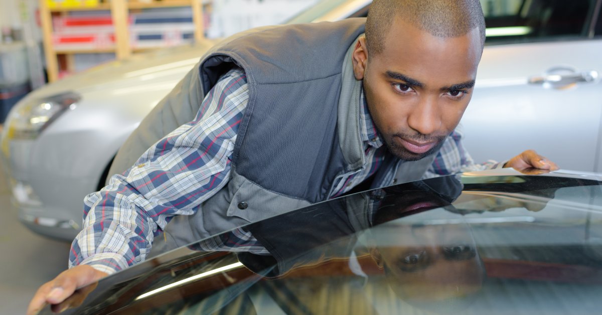 A man wearing a plaid shirt and a grey vest leans down to inspect a car's windshield. A silver car is behind him.