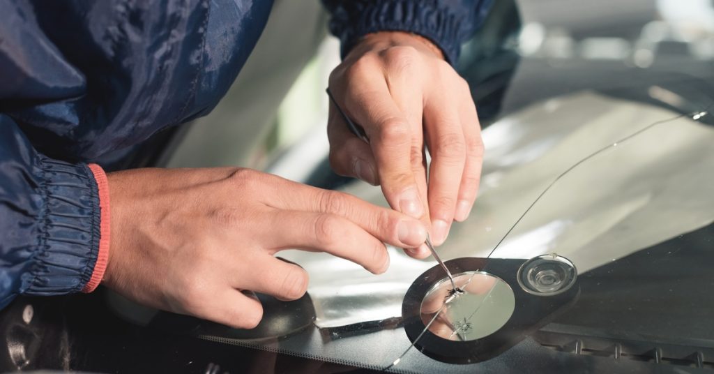An extreme close-up showing an individual wearing a blue jacket using a small tool to fix a cracked windshield.