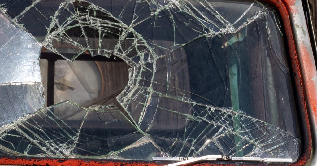 An extreme close-up showing the severely damaged and cracked windshield on a rusty, vintage red vehicle.