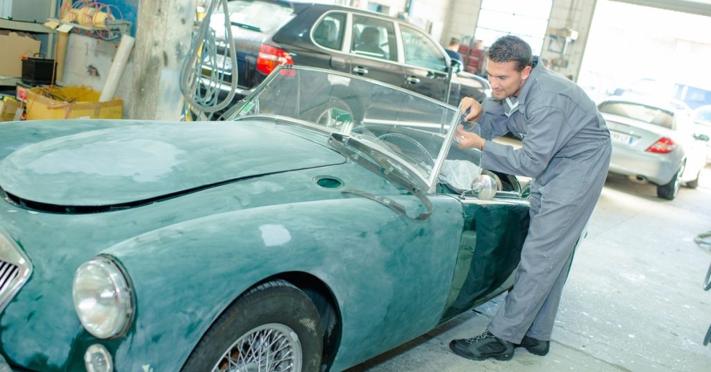A man wearing a grey jumpsuit stands inside a garage while closely inspecting the windshield on a classic car.